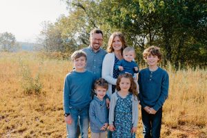 A family photo in a sunny field. All family members are wearing denim blue. The small daughter and mother each wear a light sweater over a blue outfit. The famliy has a dad, a mom, and 5 children, one of whom is a baby held by their mother.