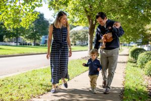 a family of four walks down a sunny sidewalk. Dad carries a baby in his arms and both parents watch as a toddler walks.