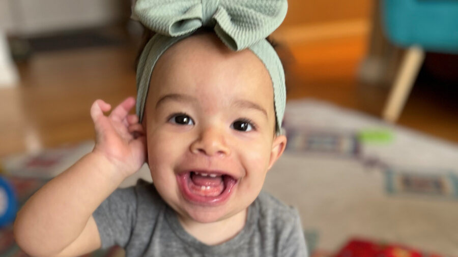 Infant girls with hair bow smiles at the camera with her mouth open and tiny teeth visible.