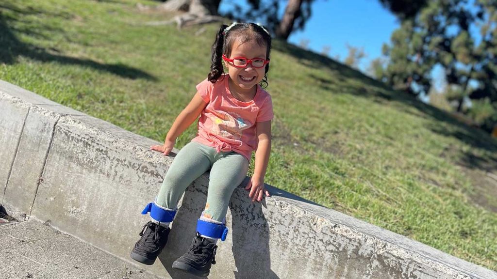 Toddler girl sits on a conrete wall with grass behind her. She's wearing red eyeglasses and smiling at the camera. SHe is also wearing leg braces.