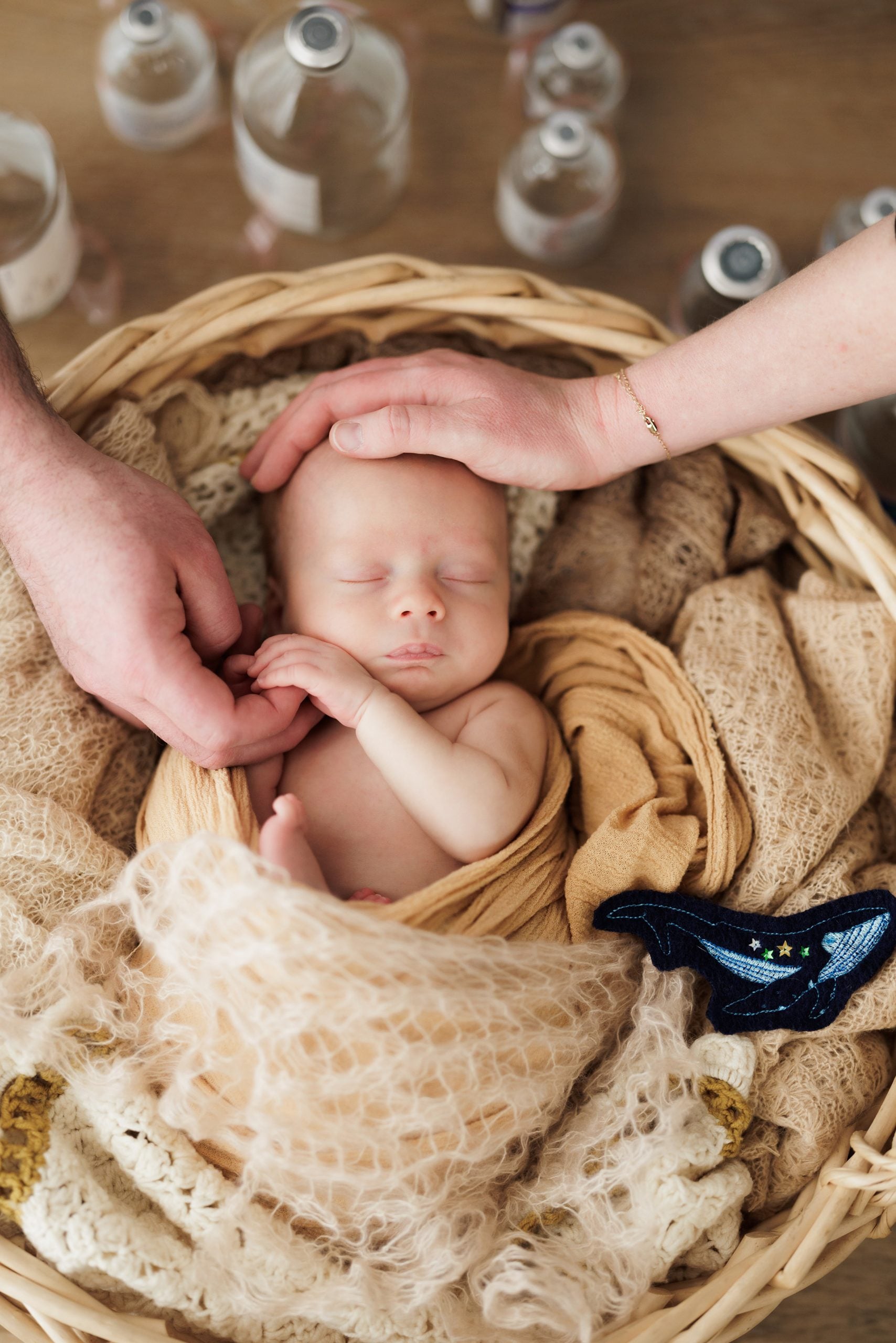 newborn baby Callum Gray photo session Baby boy peacefully sleeps in a basket swaddled in a blanket while his parents' hands gently touch him. He's surrounded by IVIG bottles and a stuffed whale to symbolize his brother, Elijah, who passed