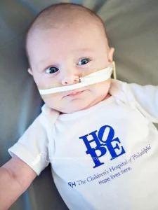 Baby boy lies in bed wearing a CHOP HOPE onesie and an oxygen tube.