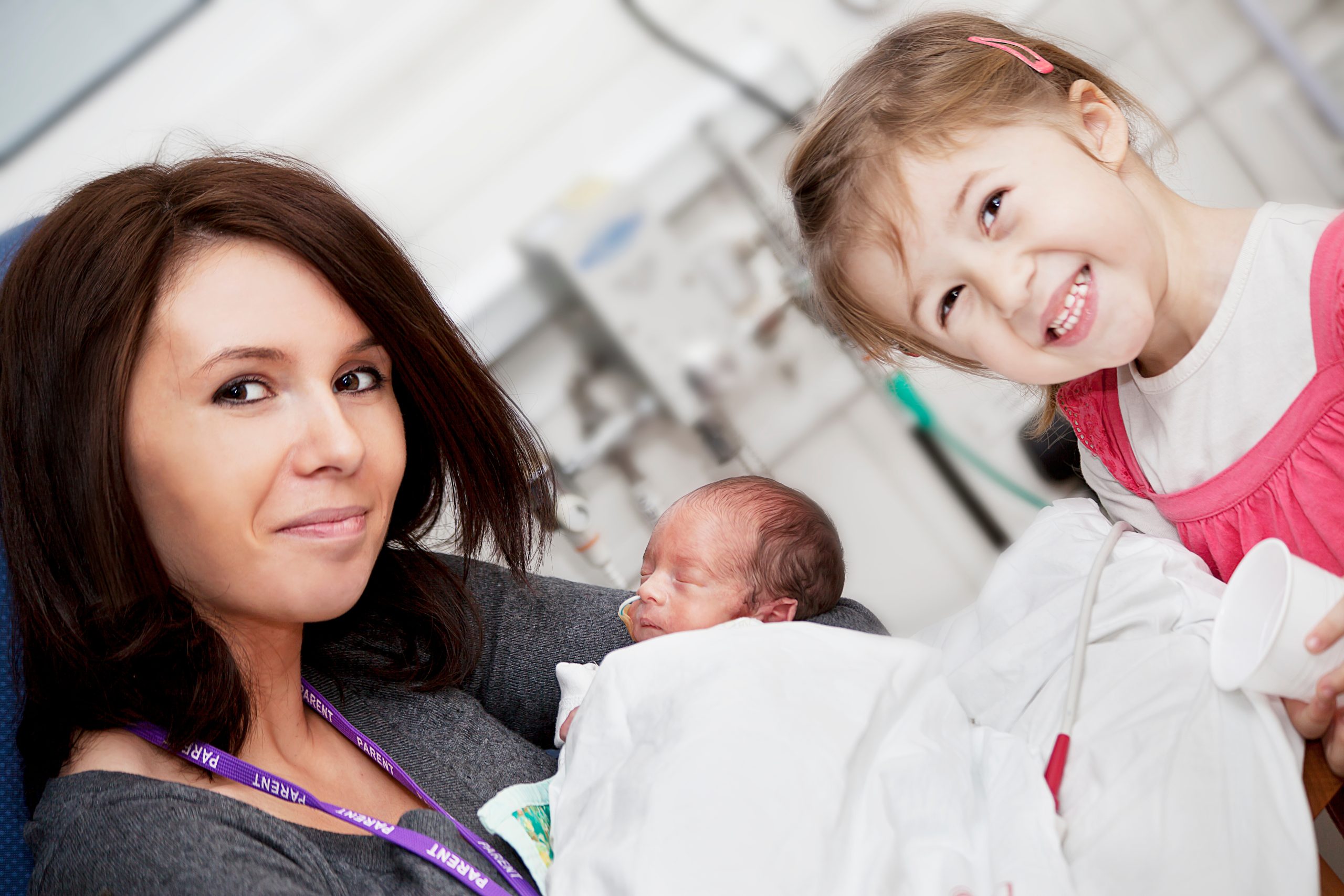 family in hospital Mom holds newborn baby in a Maternal-Fetal Surgery Center, following treatment, with toddler smiling beside them.