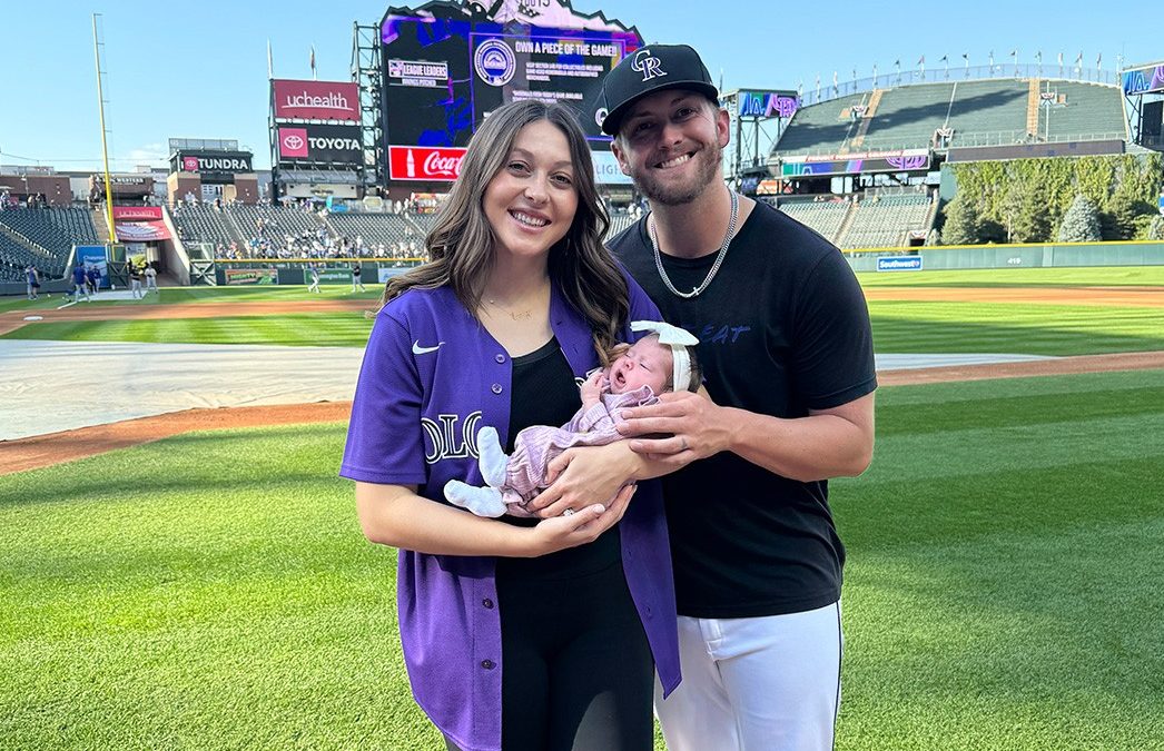 Mom and Dad hold baby Lainey on baseball field after she overcame congenital heart defects.